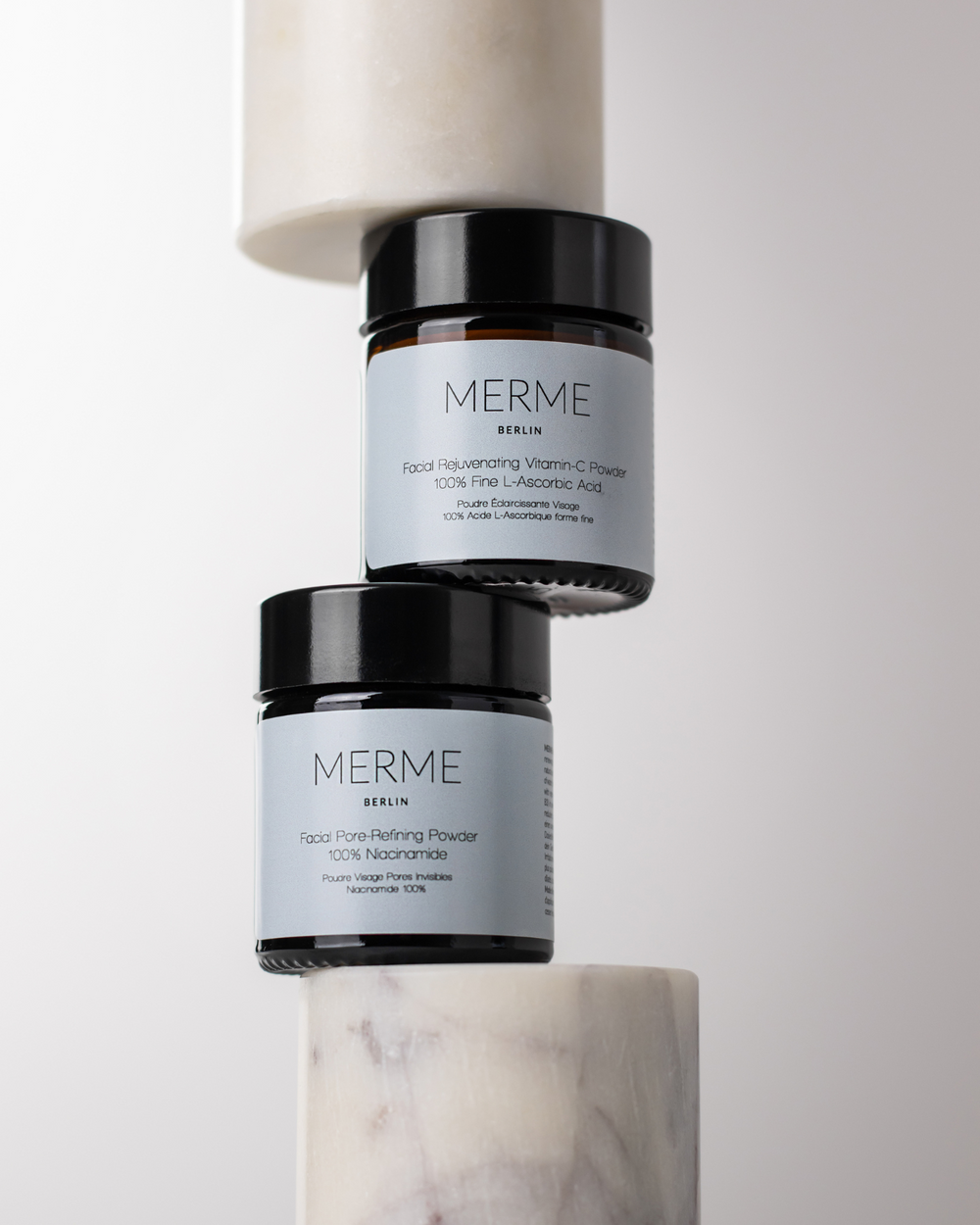 Zwei braune Glasbehälter aus dem AM/PM Sparset von MERME Berlin, einer davon Vitamin-C-Pulver und der andere Facial Pore-Refining Powder mit Niacinamid, sind vor einem minimalistischen Hintergrund auf weißen Marmorzylindern gestapelt.