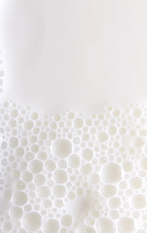Nahaufnahme von weißer Milch mit zahlreichen Blasen an der Oberfläche, die eine schaumige Textur bilden. Die Blasen variieren in ihrer Größe und bedecken die untere Bildhälfte vor einem glatten, weißen Hintergrund.