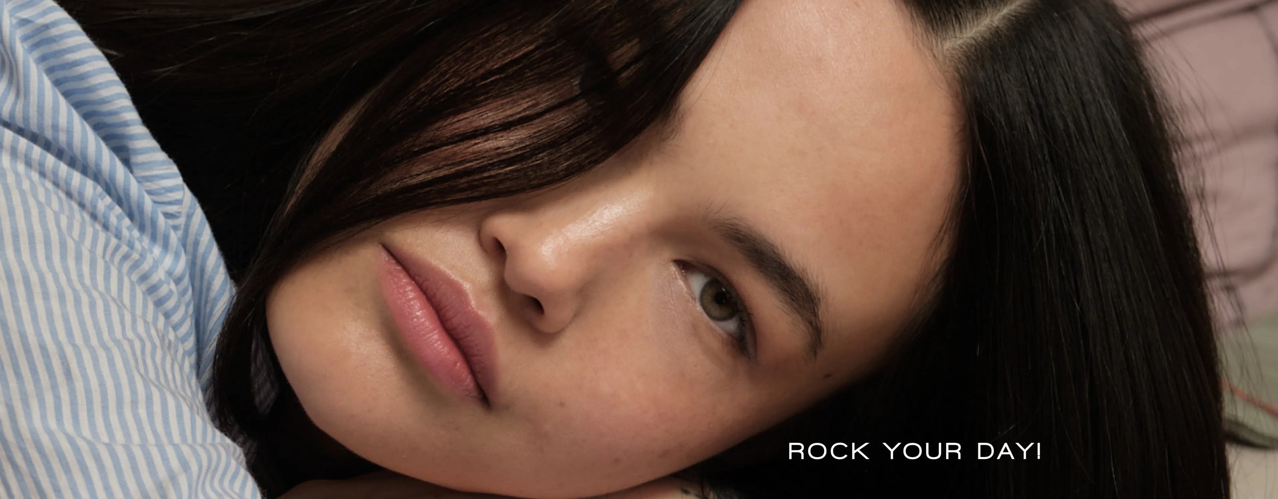 Nahaufnahme einer Frau mit glattem, dunklem Haar, die auf der Seite liegt und mit neutralem Gesichtsausdruck in die Kamera blickt. Sie trägt ein hellblau gestreiftes Hemd. Unten rechts steht: „ROCK YOUR DAY!“.