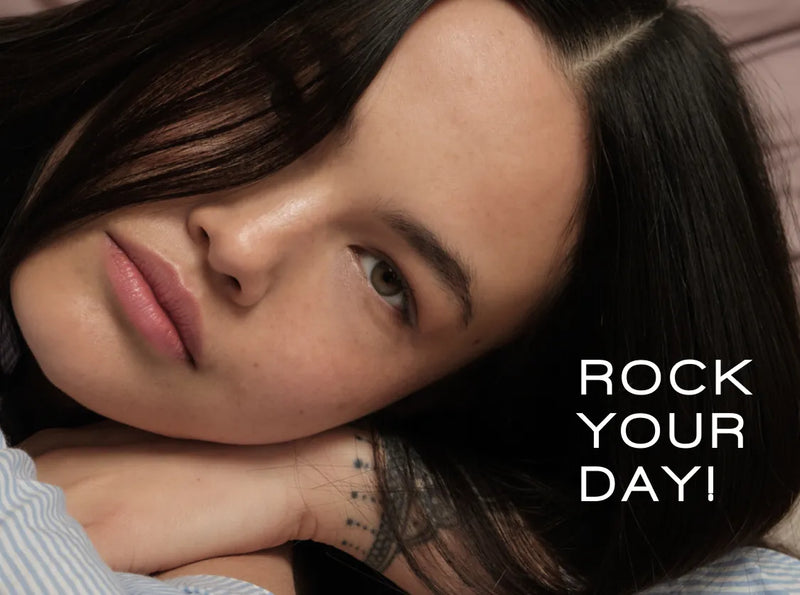 Eine Frau mit langen dunklen Haaren stützt ihren Kopf auf ihre Arme und blickt in die Kamera. Ihr Gesichtsausdruck ist neutral, und an ihrem Handgelenk ist ein Tattoo zu sehen. Neben ihr steht der Text „ROCK YOUR DAY!“.