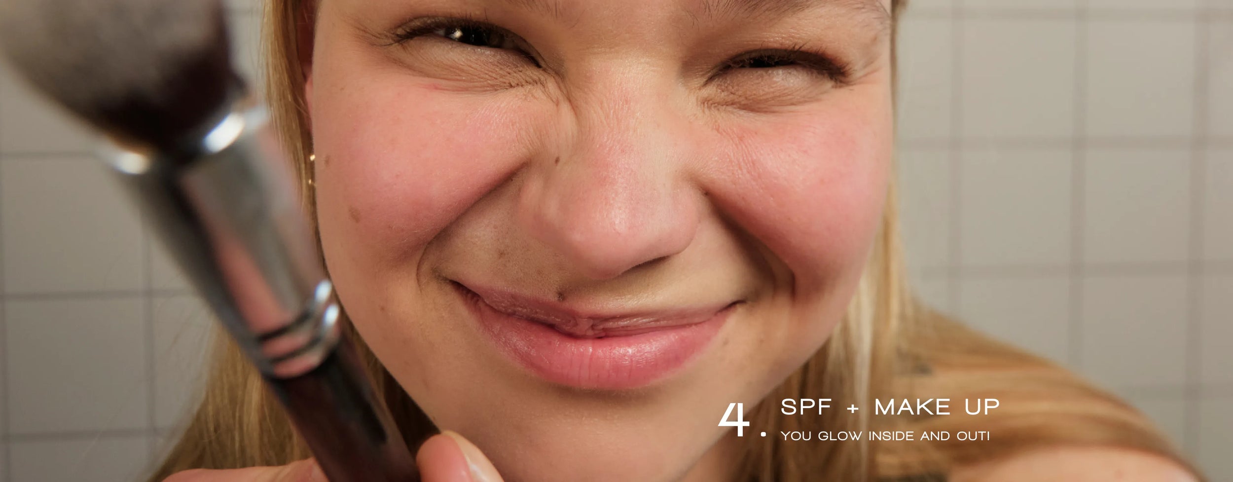 Eine lächelnde Person hält in einem hellen Badezimmer einen Make-up-Pinsel vor ihr Gesicht. Der Text lautet: „4. LSF + MAKE-UP. DU STRAHLST VON INNEN UND AUSSEN!“.