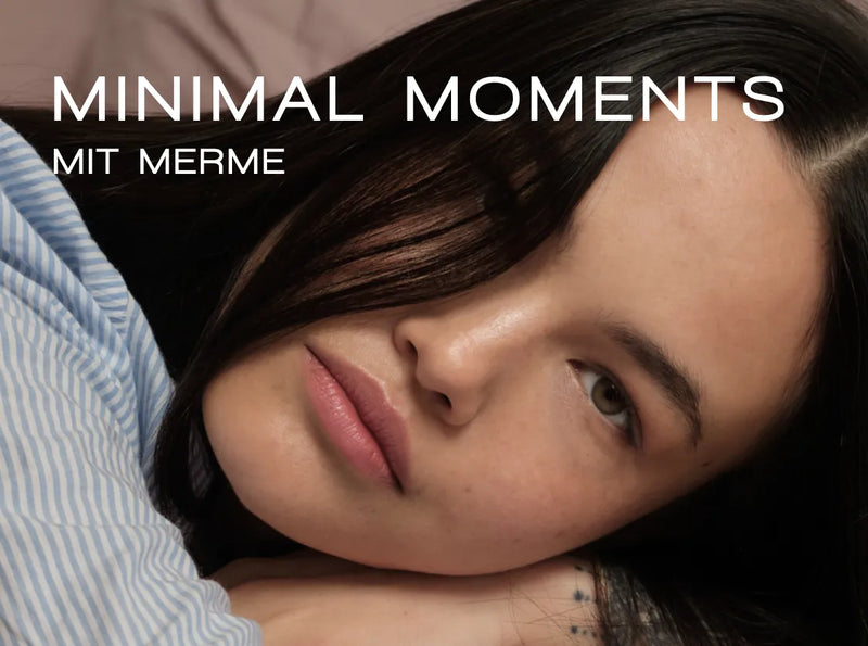 Nahaufnahme einer Person mit langen dunklen Haaren, die ihren Kopf auf den Arm stützt und ein blau gestreiftes Hemd trägt. Weißer Text über dem Bild lautet „MINIMAL MOMENTS MIT MERME“.