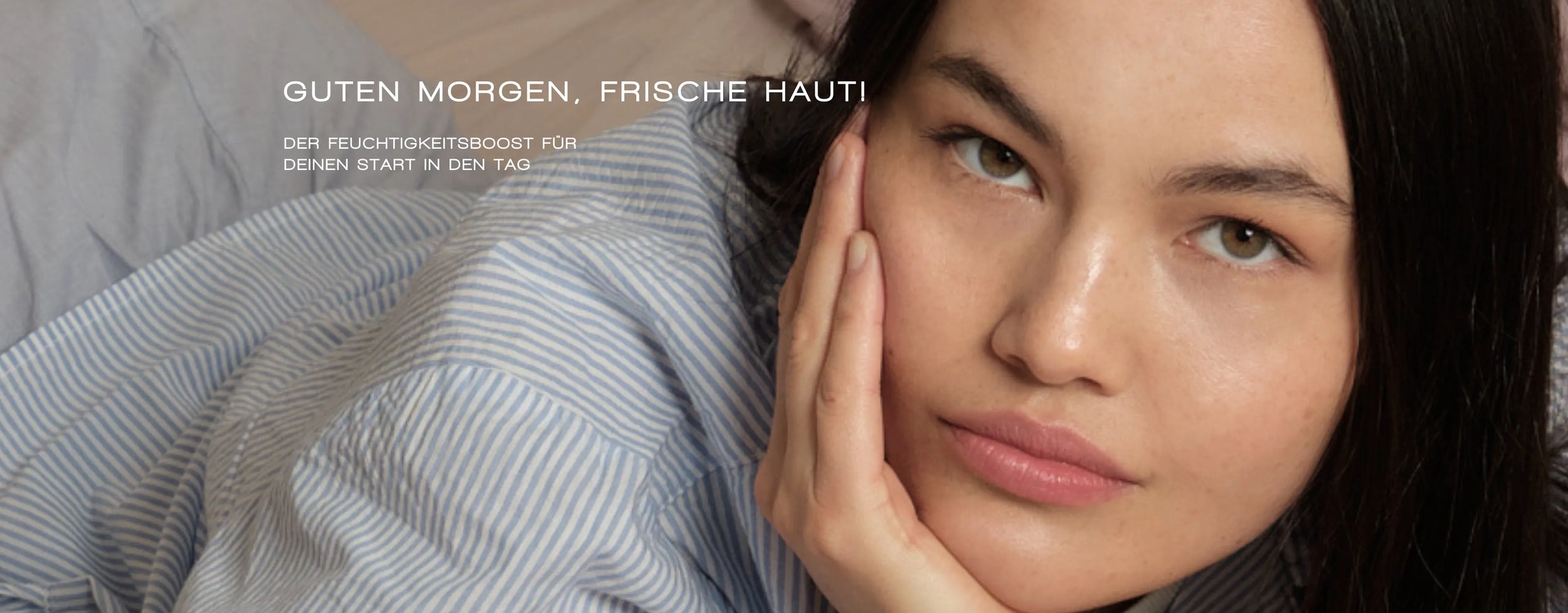 Eine Frau mit klarer Haut stützt ihr Gesicht auf ihre Hand, während sie auf einem hellen Bett liegt. Der Text auf Deutsch lautet: „Guten Morgen, frische Haut! Der Feuchtigkeitsboost für deinen Start in den Tag.“.