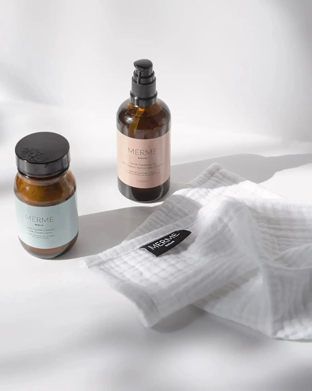 Zwei braune MERME Berlin-Flaschen - eine mit schwarzer Pumpe, eine mit schwarzem Deckel - stehen neben einem gefalteten weißen Mulltuch aus dem Set 4 x FACIAL CLEANSING CLOTHS mit der Aufschrift "MERME" auf einem weichen weißen Hintergrund mit sanften Schatten.