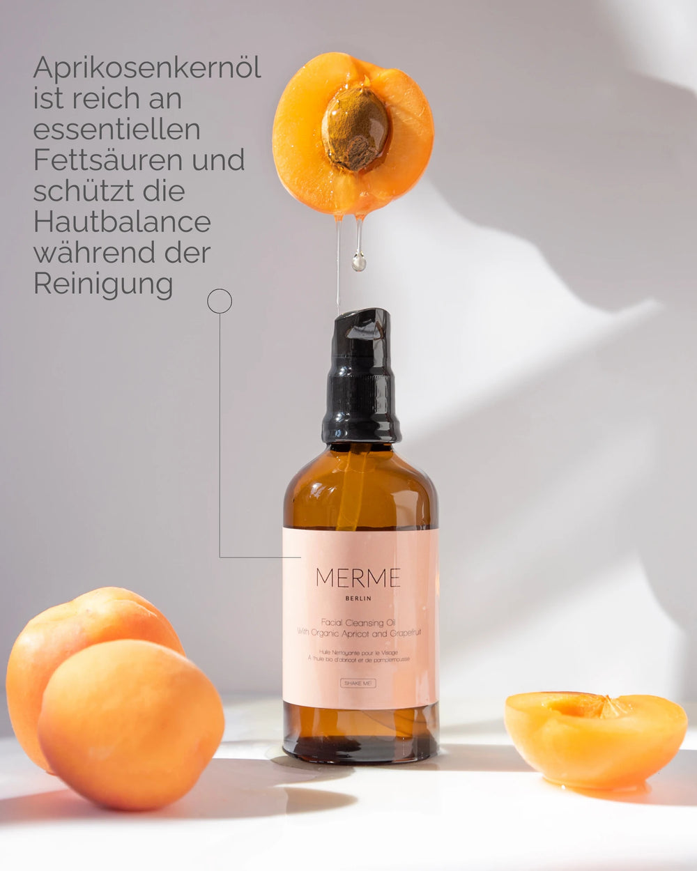 Eine Flasche MERME Berlin FACIAL CLEANSING OIL mit Grapefruit steht zwischen frischen Aprikosen, aus einer tropft Öl. Der deutsche Text erklärt, wie Bio-Aprikosenkernöl während der Reinigung hilft, die Haut auszugleichen.