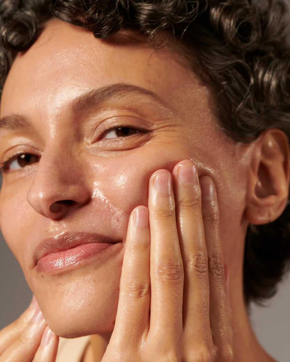 Eine Person mit strahlender Haut und lockigem Haar lächelt sanft, während sie mit den Fingern das GESICHTSREINIGUNGSÖL mit Bio-Aprikosen- und Grapefruitpackungen von MERME Berlin auf ihre Wange aufträgt.