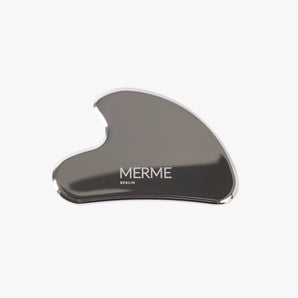 Das STAINLESS STEEL GUA SHA von MERME Berlin aus Edelstahl mit glatter, geschwungener Form und Logo-Aufdruck ist ideal zur Lymphdrainage im Gesicht und wurde vor weißem Hintergrund fotografiert.
