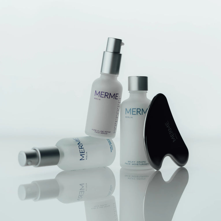 Drei Hautpflegeflaschen mit der Aufschrift „MERME Berlin“ und ein schwarzes Gua-Sha-Gerät für das Gesicht sind auf einer weißen, reflektierenden Oberfläche angeordnet und schaffen so eine minimalistische, klare Ästhetik.
