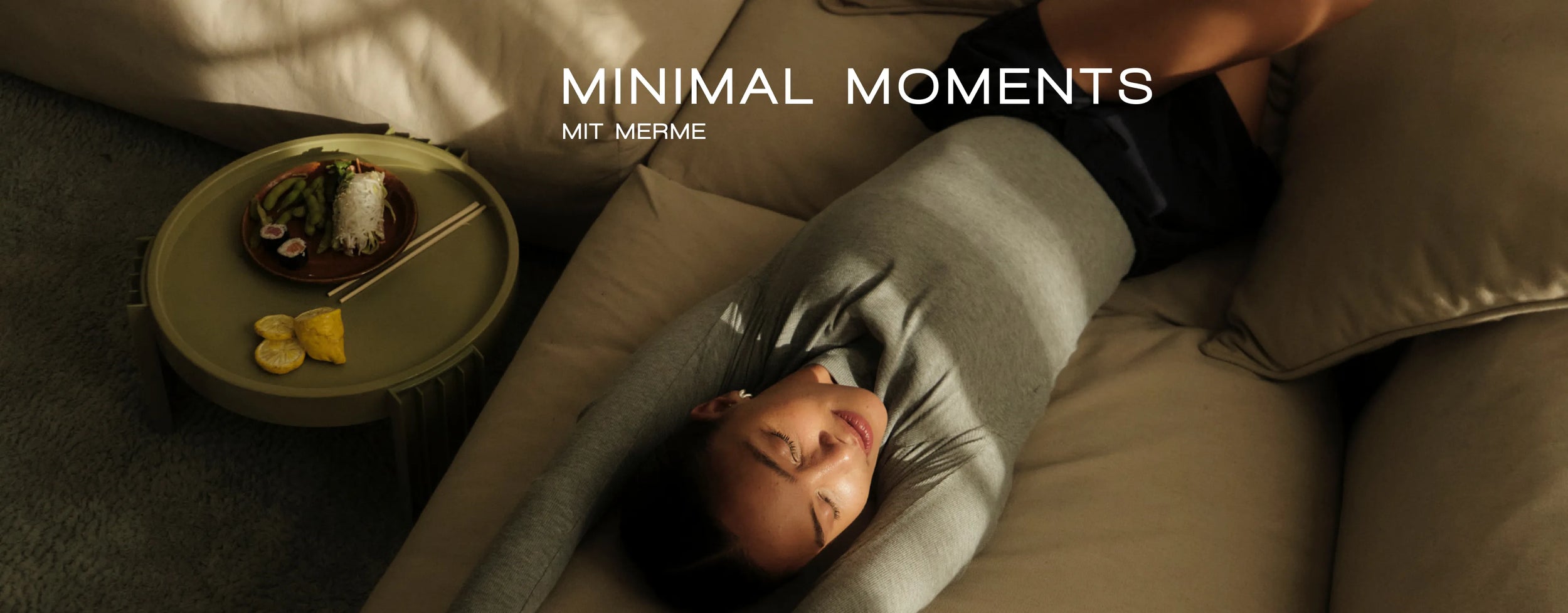 Eine Frau in einem grauen Pullover lümmelt auf einem beigen Sofa mit ausgestreckten Armen. Neben ihr steht auf einem kleinen runden Tisch ein Teller mit Essen, Stäbchen und Zitronenscheiben. Der Text lautet: „MINIMAL MOMENTS MIT MERME.“.