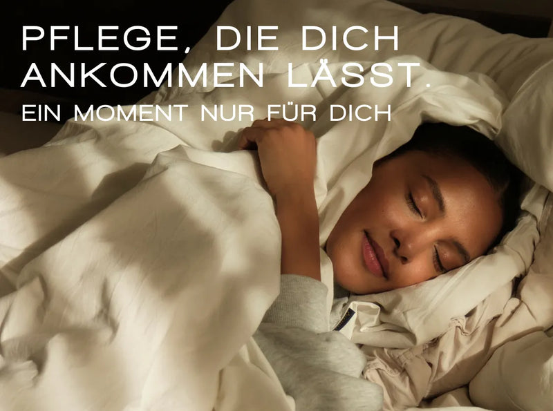 Eine Frau liegt mit einem friedlichen Lächeln bequem im Bett, in weiße Laken gehüllt. Sonnenlicht beleuchtet sanft ihr Gesicht. Der deutsche Text oben lautet: „PFLEGE, DIE DICH ANKOMMEN LÄSST. EIN MOMENT NUR FÜR DICH.“.
