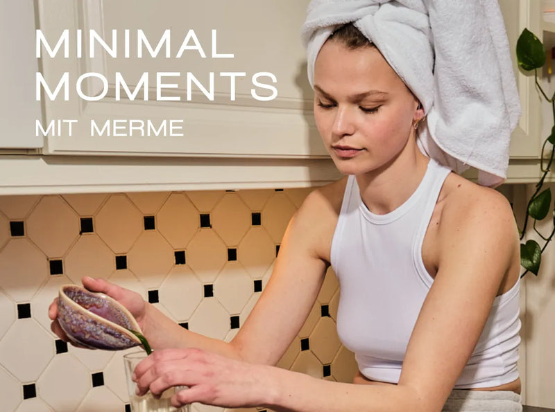 Eine Frau mit einem Handtuch um den Kopf und einem weißen Tanktop presst in einer Küche eine Zitrone in ein Glas. Der Text auf dem Bild lautet: „MINIMAL MOMENTS MIT MERME.“.
