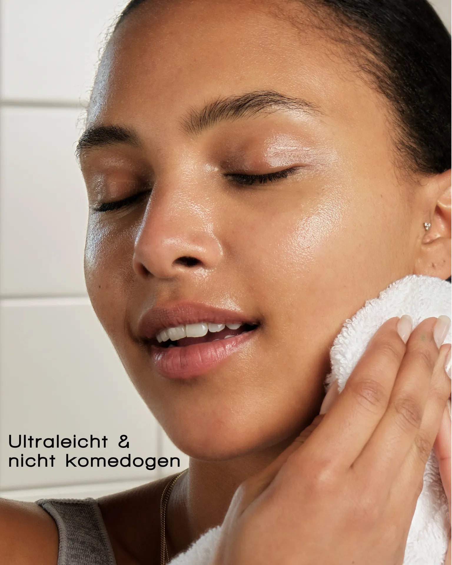 Eine Frau mit leuchtender Haut lächelt sanft und drückt ein weißes Handtuch an ihr Gesicht. Der Text lautet: "Ultraleicht & nicht komedogen"-DELICATE DISSOLVE FACE CLEANSER von MERME Berlin, perfekt für die sanfte Reinigung und empfindliche Haut.