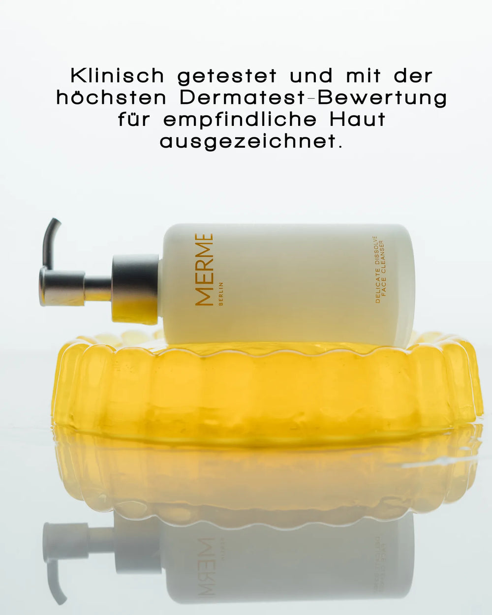 Eine Flasche MERME Berlin DELICATE DISSOLVE FACE CLEANSER mit einer Pumpe steht auf einer gelben, durchsichtigen Seifenschale. Oben weist ein deutscher Text auf die klinischen Tests und die beste Dermatest-Bewertung für empfindliche Haut hin. Der Hintergrund ist weiß.