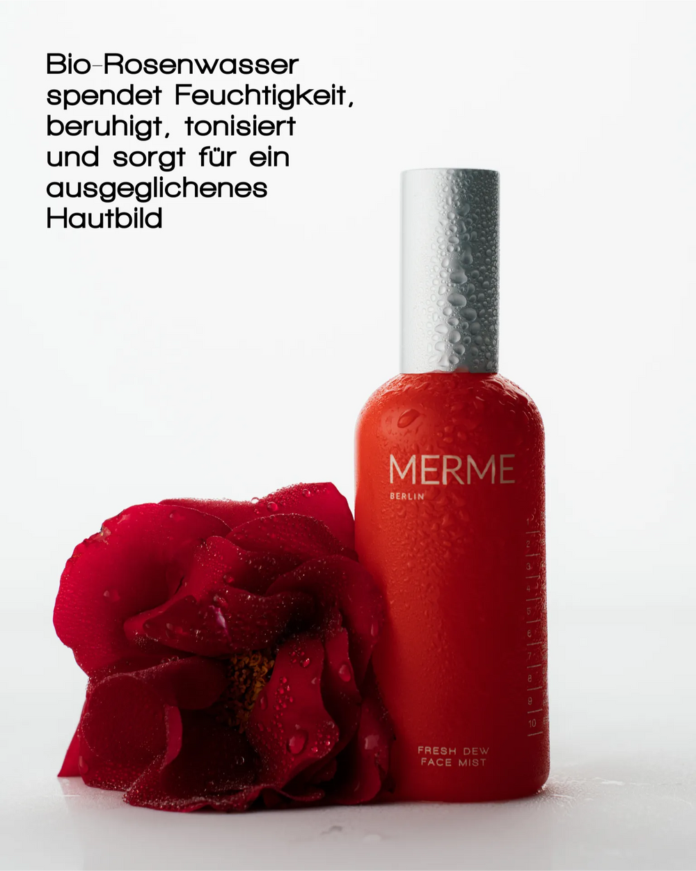 Ein roter Flakon von MERME Berlin FRESH DEW FACE MIST mit Wassertropfen steht neben einer taufrischen roten Rose auf weißem Hintergrund. Der deutsche Text unterstreicht die Vorteile von MERME FRESH DEW FACE MIST, das die Haut mit Feuchtigkeit versorgt und ihr ein strahlendes Aussehen verleiht.