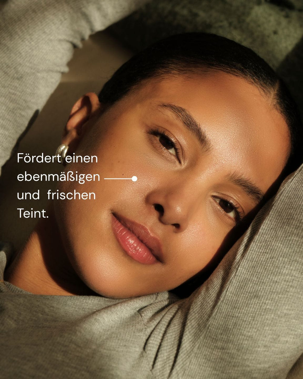 Eine Frau mit strahlender Haut liegt mit erhobenen Armen und blickt in die Kamera. Der deutsche Text lautet: "Fördert einen ebenmäßigen und frischen Teint - für natürliche Ausstrahlung." GLOWING GUARD FACE OIL von MERME Berlin.