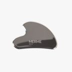 Die Edelstahl Gua Sha von MERME Berlin mit gebogener Form und Logo ist auf weißem Hintergrund zu sehen - das ideale Massage-Tool für entspannende Lymphdrainage.
