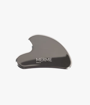 Die Edelstahl Gua Sha von MERME Berlin mit gebogener Form und Logo ist auf weißem Hintergrund zu sehen - das ideale Massage-Tool für entspannende Lymphdrainage.
