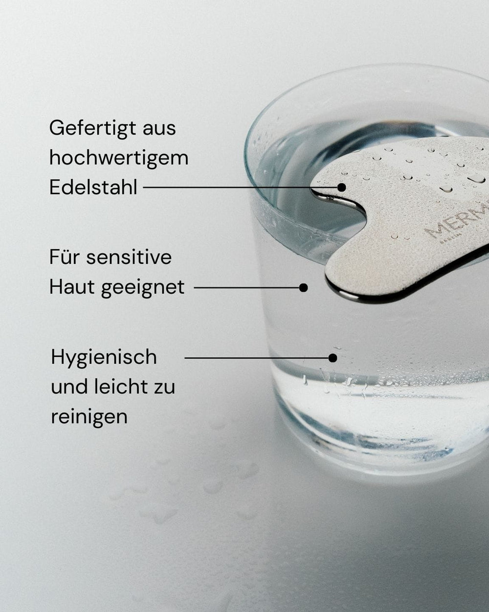 Das MERME Berlin Edelstahl Gua Sha liegt auf dem Glasrand. Der deutsche Text betont: ideal für Gesichtsmassage & Lymphdrainage, aus hochwertigem Edelstahl, hygienisch, leicht zu reinigen und auch für empfindliche Haut geeignet.