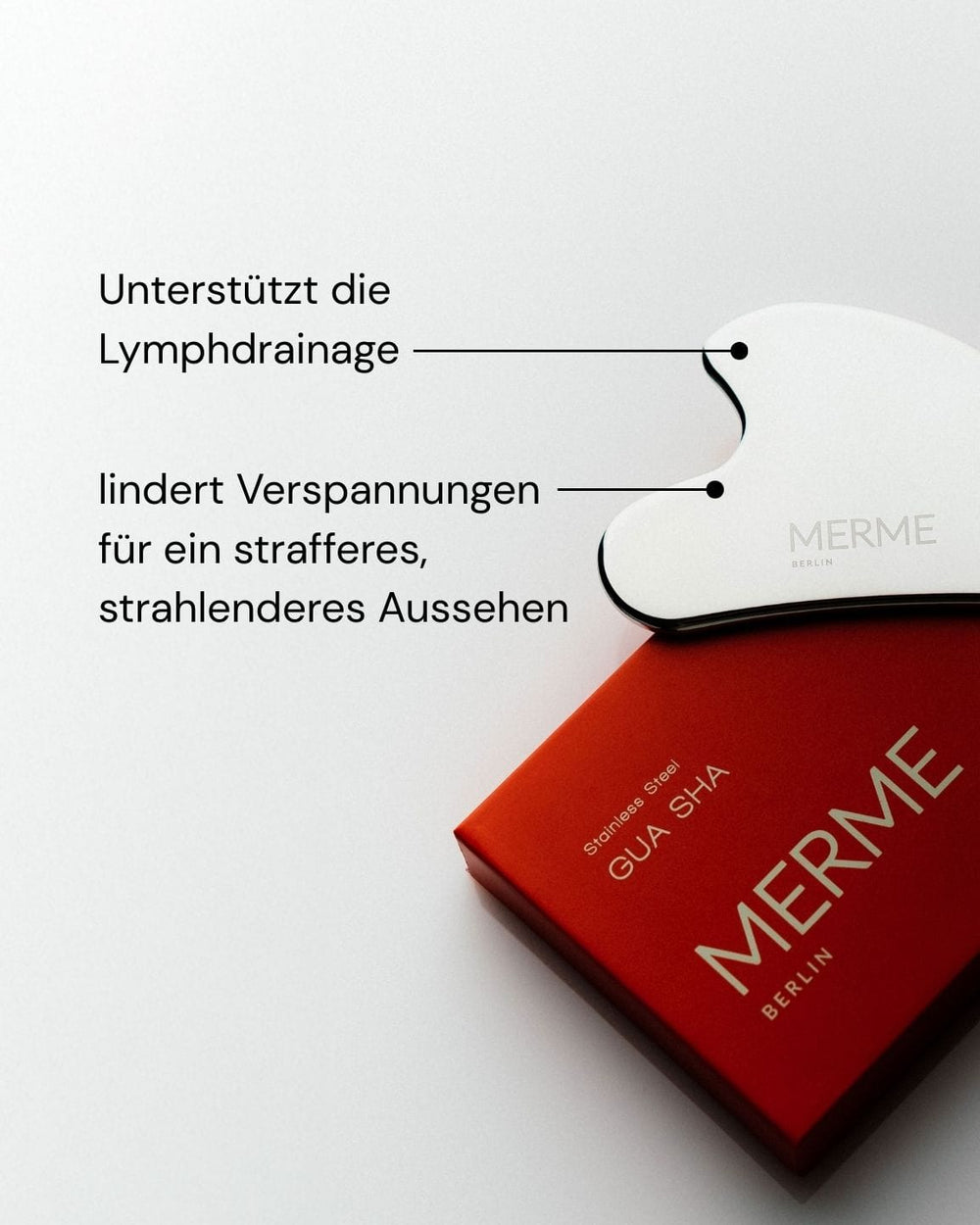 Ein weißes Gua Sha aus Edelstahl von MERME Berlin ruht auf einer roten Schachtel. Schwarze Linien und deutscher Text heben seine Vorzüge hervor: "fördert die Lymphdrainage und Gesichtsmassage für straffere, strahlendere Haut".