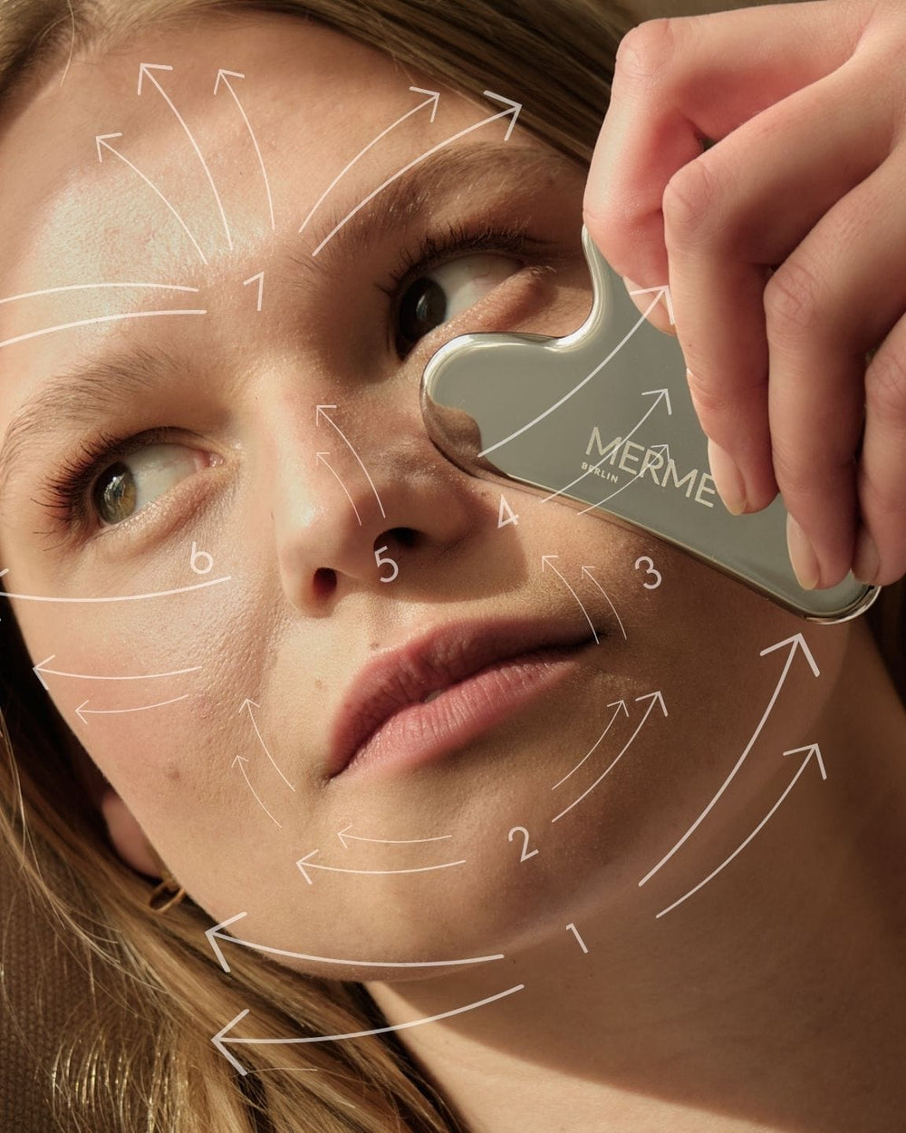 Eine Frau verwendet das MERME Berlin Edelstahl Gua Sha für eine Gesichtsmassage an ihrer Wange. Weiße Pfeile und Zahlen markieren die Massagerichtung und betonen die Lymphdrainagetechniken für eine effektive Hautpflege.