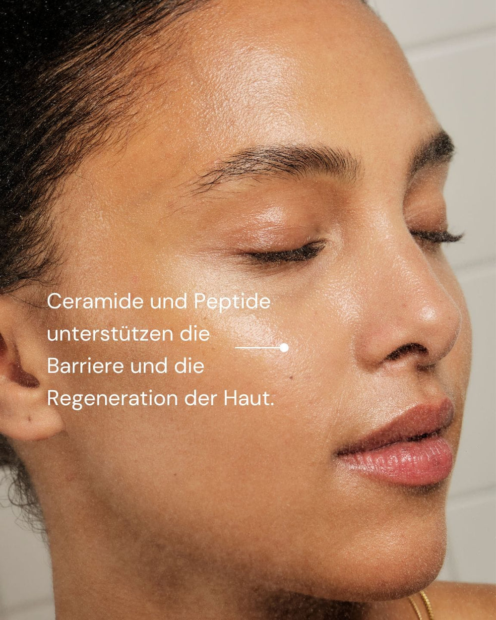 Eine Nahaufnahme der strahlenden Haut einer Frau. Deutscher Text: "Ceramide und Peptide unterstützen die Barriere und Regeneration der Haut." MERME Berlin Milky Drops Face Moisturiser-perfekte Feuchtigkeitspflege vor neutralem hellem Hintergrund.