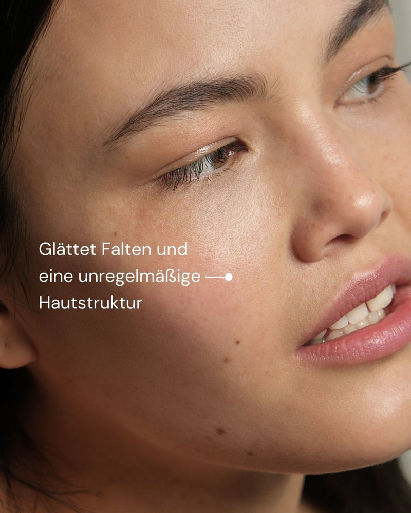 Eine Nahaufnahme der glatten, klaren Haut einer Frau mit deutschem Text: "Glättet Falten und eine unregelmäßige Hautstruktur", um das New by Morning Retinal Face Serum von MERME Berlin vorzustellen, das Falten und eine unregelmäßige Hautstruktur glättet.