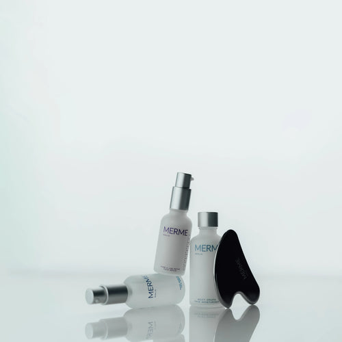 Drei MERME-Hautpflegeflaschen und ein schwarzes Gesichtsmassagegerät sind auf einer weißen reflektierenden Oberfläche mit einem minimalistischen, hellen Hintergrund angeordnet.