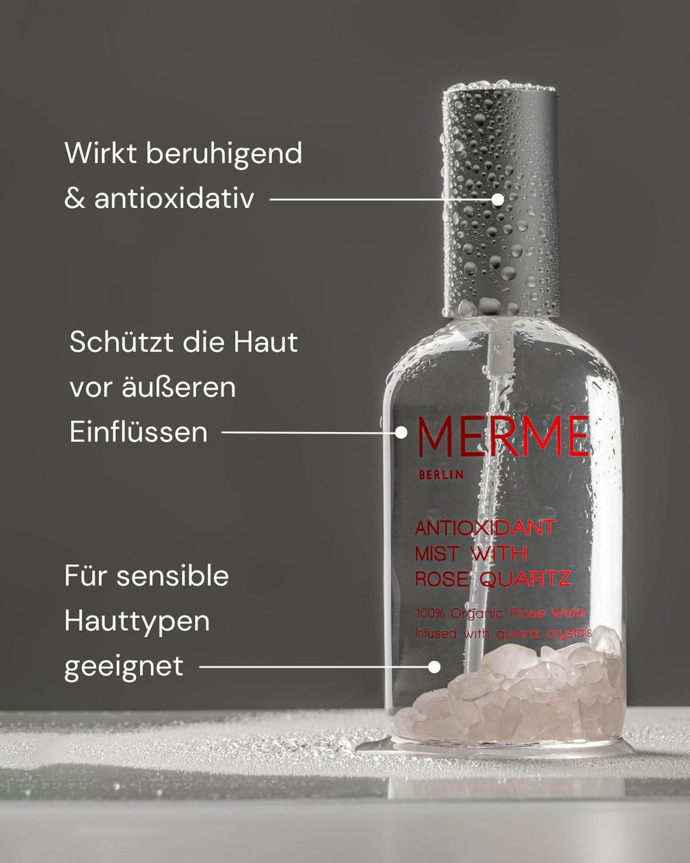 Eine klare Sprühflasche mit der Aufschrift "ANTIOX MIST WITH ROSE QUARTZ" von MERME Berlin steht auf einer grauen Fläche. Rosenquarzsplitter spenden Feuchtigkeit; der deutsche Text betont die beruhigende, antioxidative Pflege für empfindliche Haut.