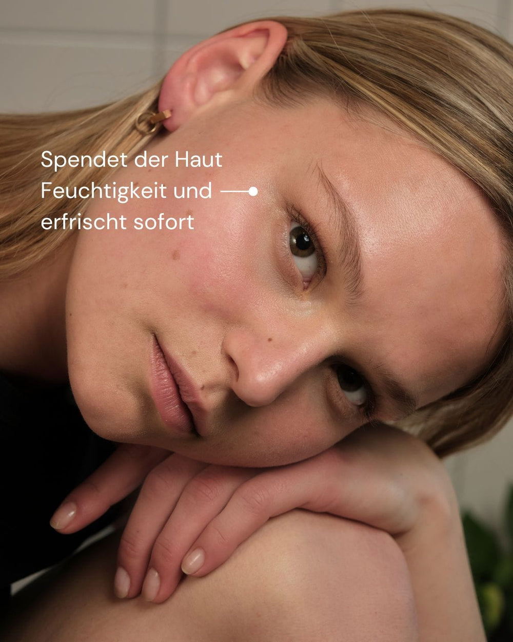 Eine Frau mit klarer, leuchtender Haut und hellbraunem Haar stützt ihren Kopf auf ihre Hände und blickt in die Kamera. Der deutsche Text lautet: "Spendet Feuchtigkeit und erfrischt sofort - ANTIOX MIST WITH ROSE QUARTZ von MERME Berlin.