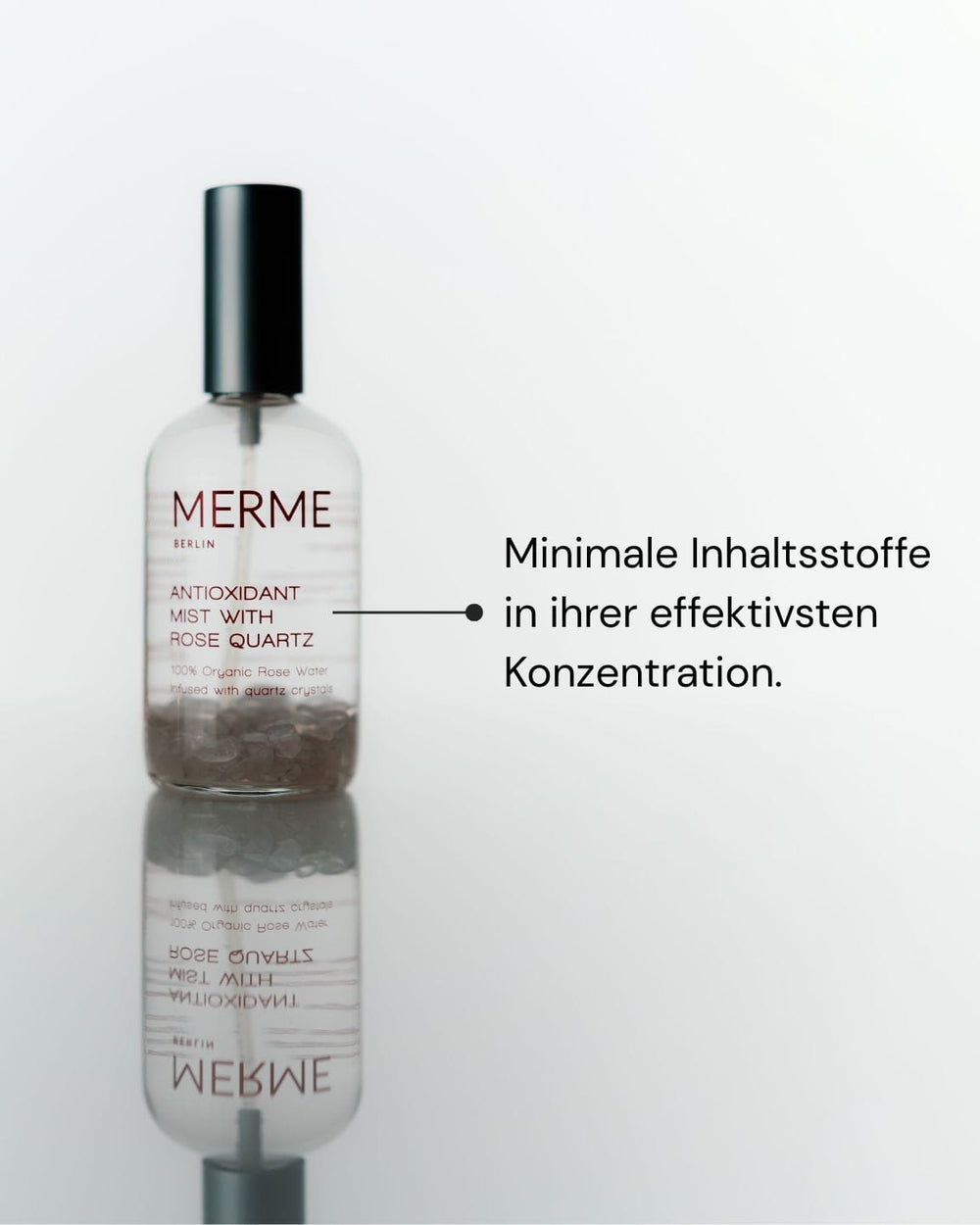 Eine Flasche MERME Berlin ANTIOXIDANT MIST WITH ROSE QUARTZ steht auf einer spiegelnden Fläche. Die Beschriftung: "Minimale Inhaltsstoffe in effektiver Konzentration - für maximale Feuchtigkeit.