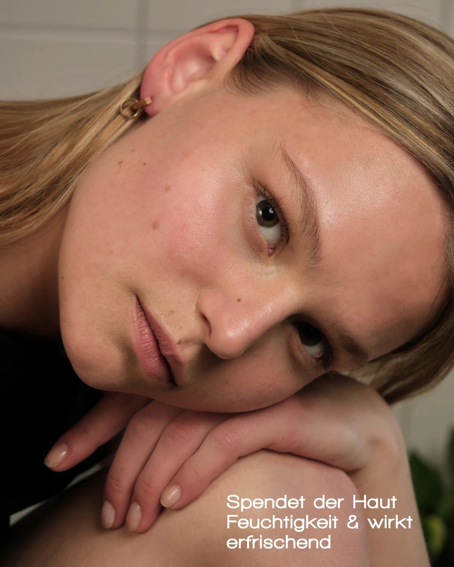 Eine Nahaufnahme einer jungen Frau mit klarer, taufrischer Haut, die ihr Kinn auf ihre Hand stützt und in die Kamera blickt. Deutscher Text: „MERME Berlin ANTIOXIDANT MIST WITH ROSE QUARTZ spendet Feuchtigkeit & erfrischt die Haut.“.
