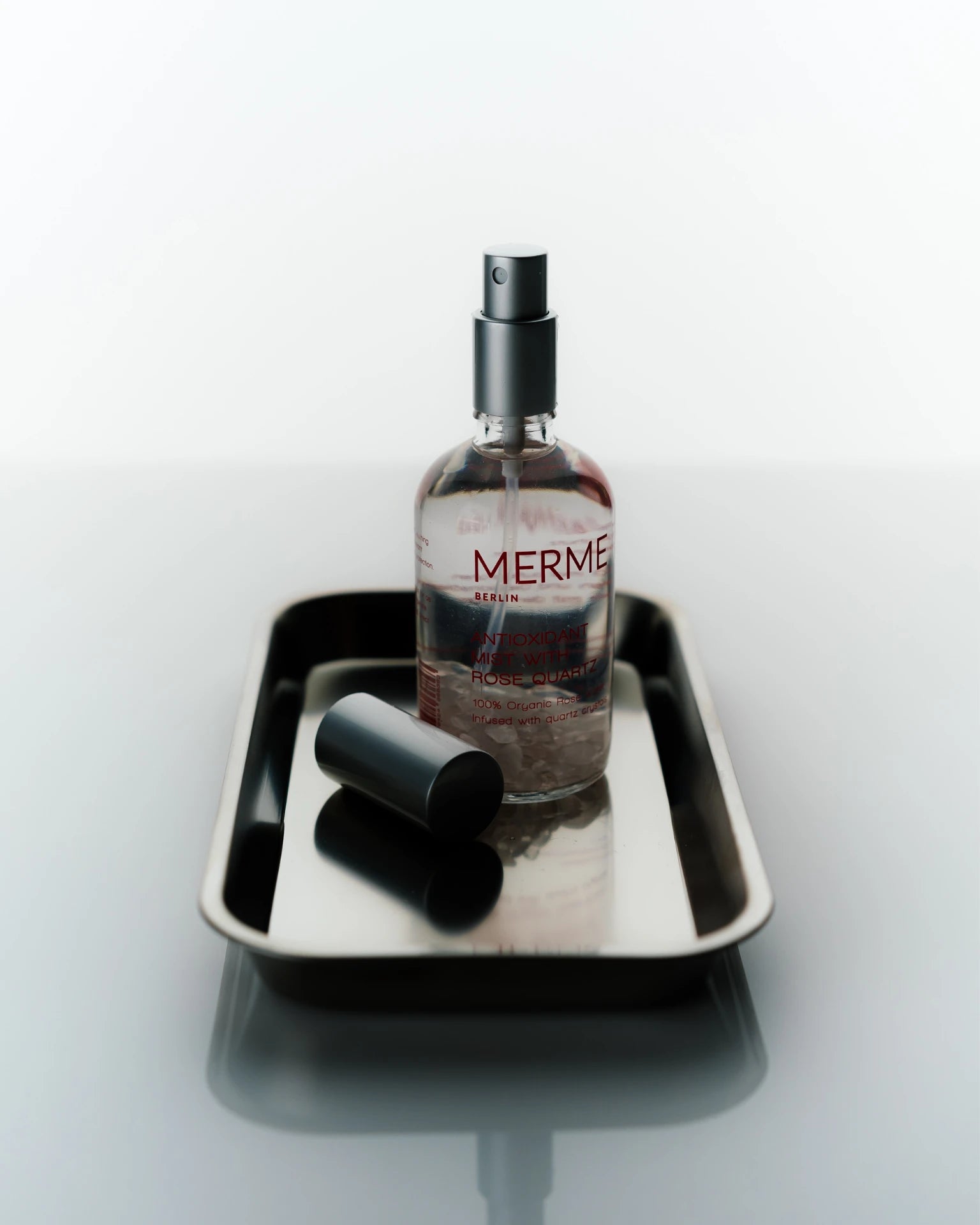 Eine Flasche MERME Berlin ANTIOXIDANT MIST WITH ROSE QUARTZ mit Sprühdüse steht auf einem rechteckigen Metalltablett; der Deckel liegt daneben und die minimalistische, reflektierende Anordnung spiegelt die Reinheit des Rosenquarzes wider.