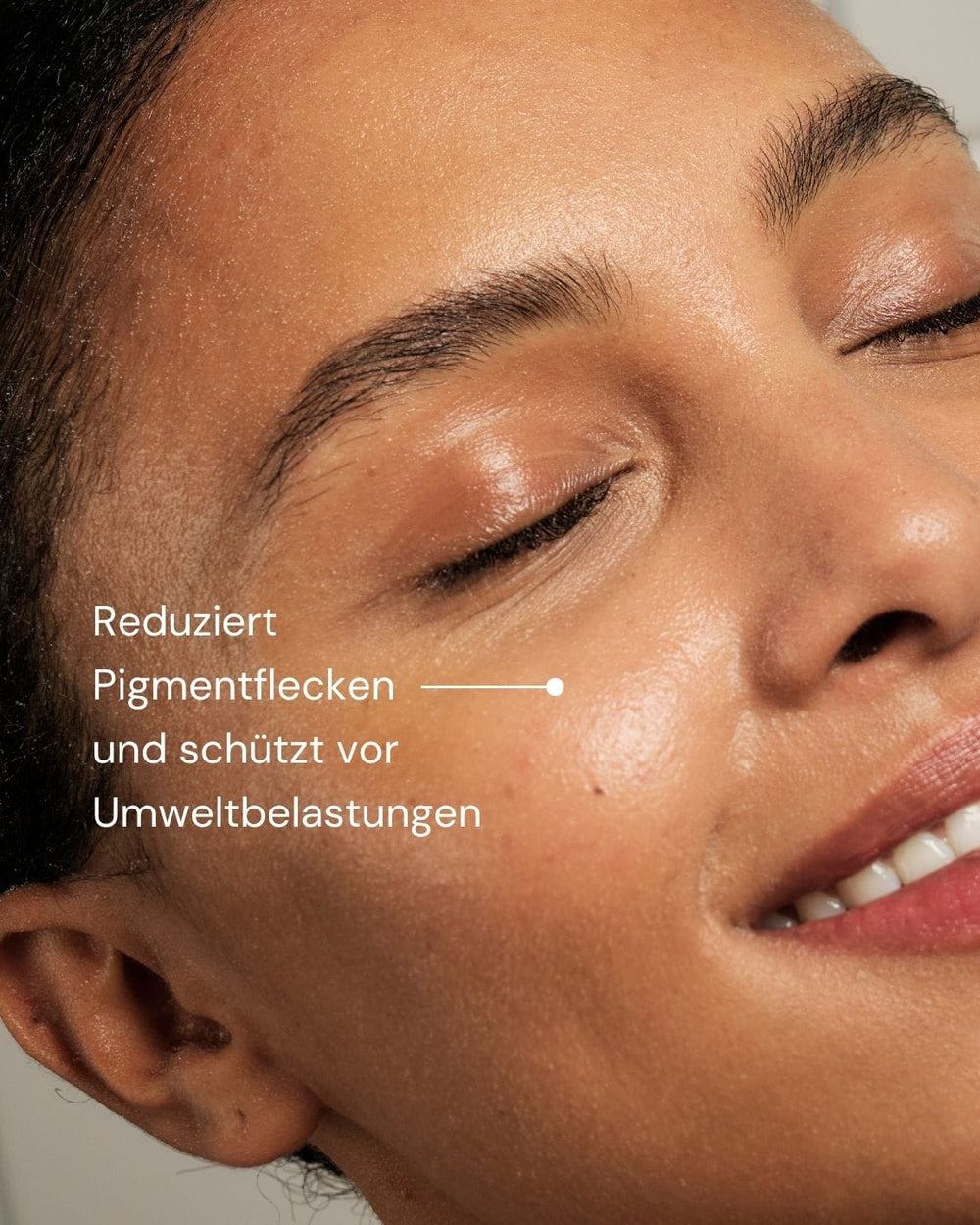 Eine Frau mit leuchtender Haut lächelt mit geschlossenen Augen vor einem gekachelten Hintergrund. Weißer Text in deutscher Sprache lautet: "Super Stable C Face Serum von MERME Berlin - reduziert Pigmentflecken und schützt vor Umweltbelastungen", mit einer Linie, die auf ihre Wange zeigt.